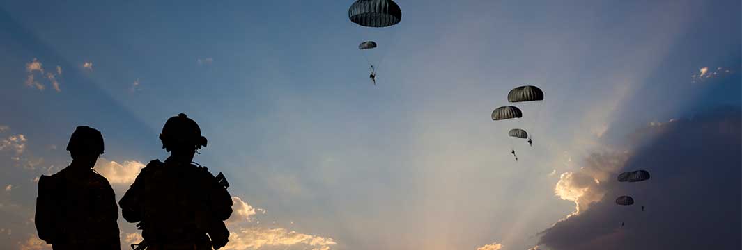 Silhouetted soldiers watch as multiple paratroopers descend under parachutes from military aircraft against a dramatic sunset sky
