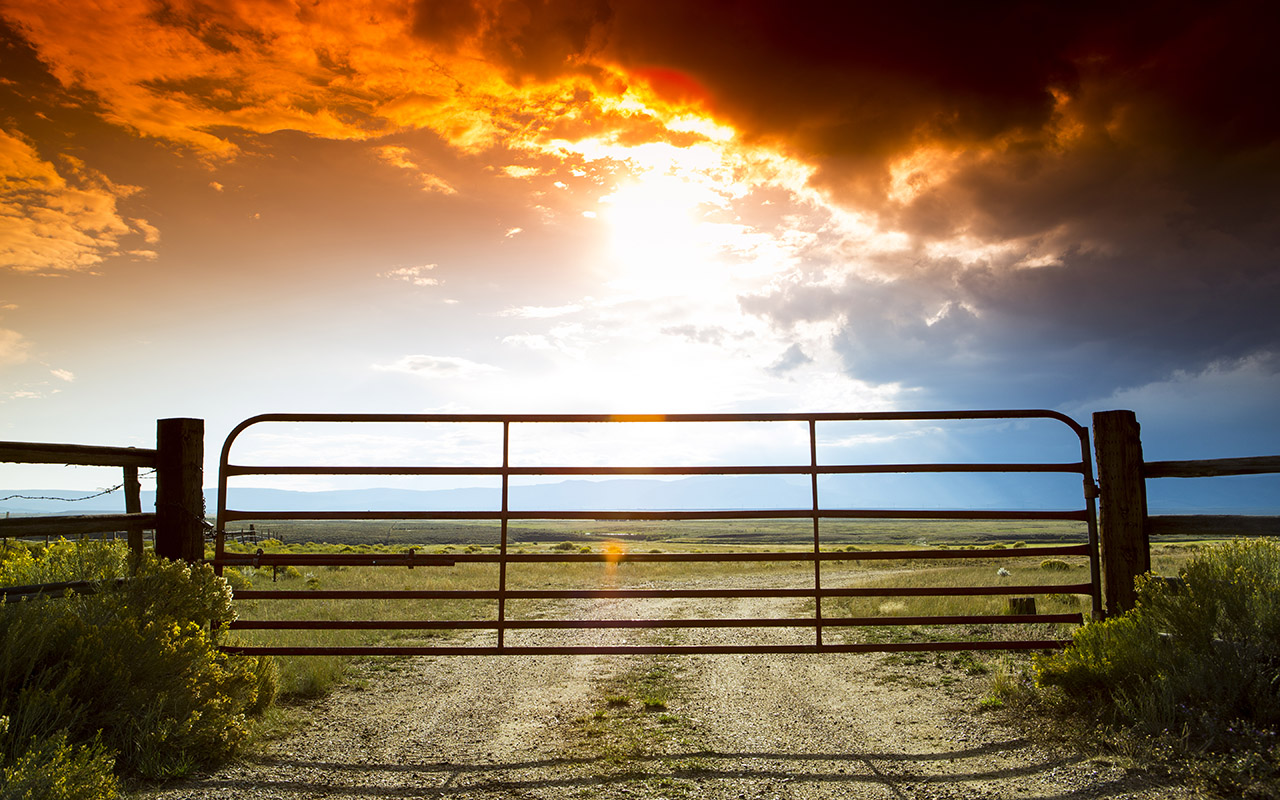 A farm fence gate with a setting sun peeking through clouds behind it.