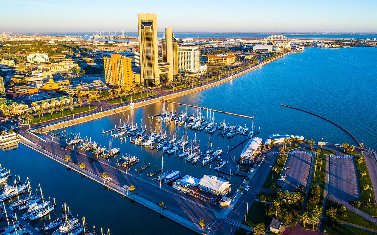 aerial photo of Paradise palm Trees and golden towers along Corpus Christi , TX Bayfront at sunrise with marina and the water still with a reflecting cityscape painted in golden hour , Marina , boats , and Bay with Gulf of mexico and sea wall . The refineries and in the background and thousands of new Wind Turbines line the background.