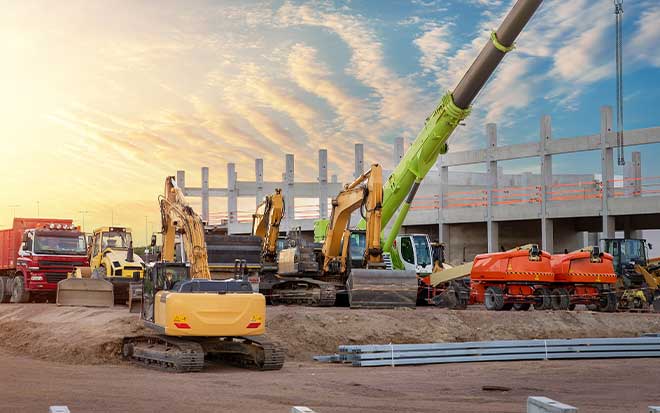 A construction site, setting sun, and early stages of a building are pictured with a crane and construction equipment.