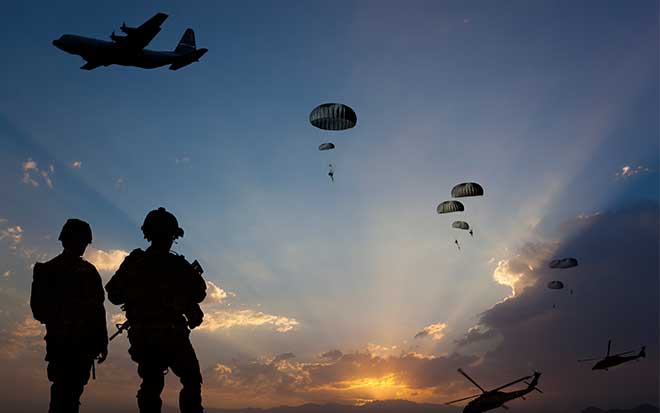 Silhouettes of soldiers watching paratroopers descend from aircraft at sunset, with multiple planes flying overhead